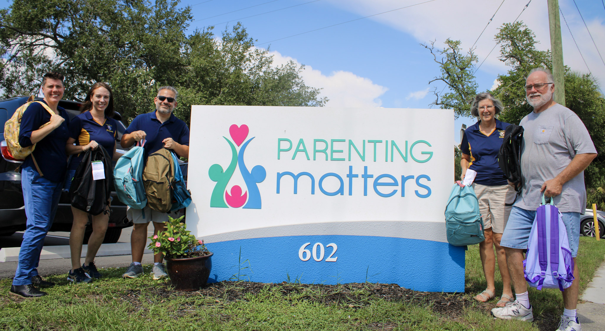 Manatee Sunrise Kiwanis Club leadership stand with Parenting Matters Executive Director Katie Becker next to the Parenting Matters sign holding backpacks